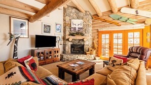 Ein gemütliches Wohnzimmer mit Kamin aus Stein, Holzbalken, einem Ledersessel und einem Fernseher.