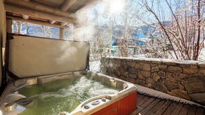 Ein Whirlpool mit Steinwand und Holzterrasse, umgeben von Bäumen und Blick auf Gebäude.