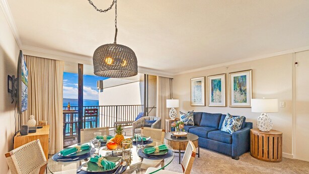 A modern living room with a glass dining table, blue sofa, and artwork on the walls. - Ocean view 2-bedroom Condo in Lahaina, West Maui - Kaanapali Shores 903 (Lahaina)