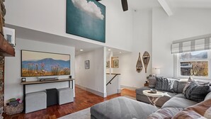 Ein modernes Wohnzimmer mit einem großen Flachbildfernseher, grauen Sofas, einem kleinen Beistelltisch und einer Treppe, die in ein Obergeschoss führt.