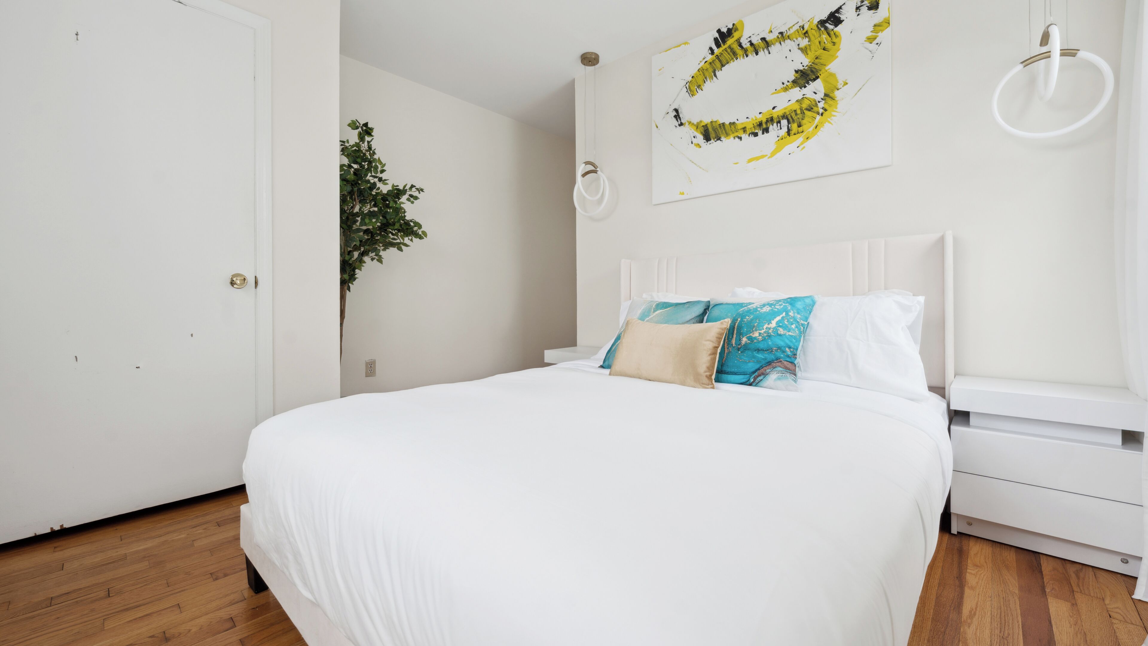 Un dormitorio con cama blanca, mesita de noche, lámpara de pared y un cuadro.