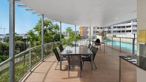 A modern outdoor terrace with a glass table and chairs, a barbecue grill, and a pool in the background. - 2-bedroom Apartment 7 km from Brisbane Airport (Chermside West)