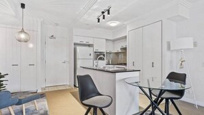 A modern kitchen with white cabinetry, a central island with a sink, and a dining area with a glass table and black chairs. - 2-bedroom Apartment 7 km from Brisbane Airport (Chermside West)
