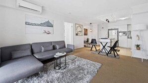 A modern living room with a grey sofa, a small round table, and a coffee table. There is a dining area with a white table and chairs, and a kitchen in the background. - 2-bedroom Apartment 7 km from Brisbane Airport (Chermside West)