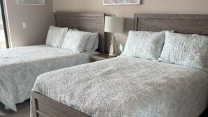 Een hotelkamer met twee bedden, een houten hoofdeinde, een nachtkastje met een lamp en een schilderij met een strandthema aan de muur.