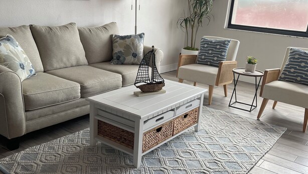 Een moderne woonkamer met een beige bank, een witte bijzettafel met manden, een kamerkleed met patroon en een groot raam met uitzicht.