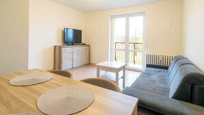 Ein modernes Wohnzimmer mit einem Holzesstisch, zwei gewebten Platzsets, einem grauen Sofa, einem Fernseher auf einem Schrank und einem großen Fenster mit Blick ins Freie.
