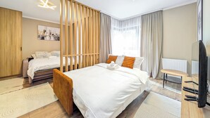 Moderni hotellihuone, jossa on suuri sänky, puinen pääty, työpöytä tuolilla, televisio ja ikkuna, jossa on verhot.
