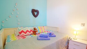 A neatly made bed with a quilt, pillows, and a stuffed toy. There is a heart-shaped decoration on the wall and a lamp on the bedside table.