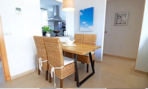 A dining area with a wooden table and wicker chairs, a pendant light, a wall-mounted picture, and a kitchen in the background. - Wake up to the waves in this beachfront Torrox apartment. (Torrox Costa)