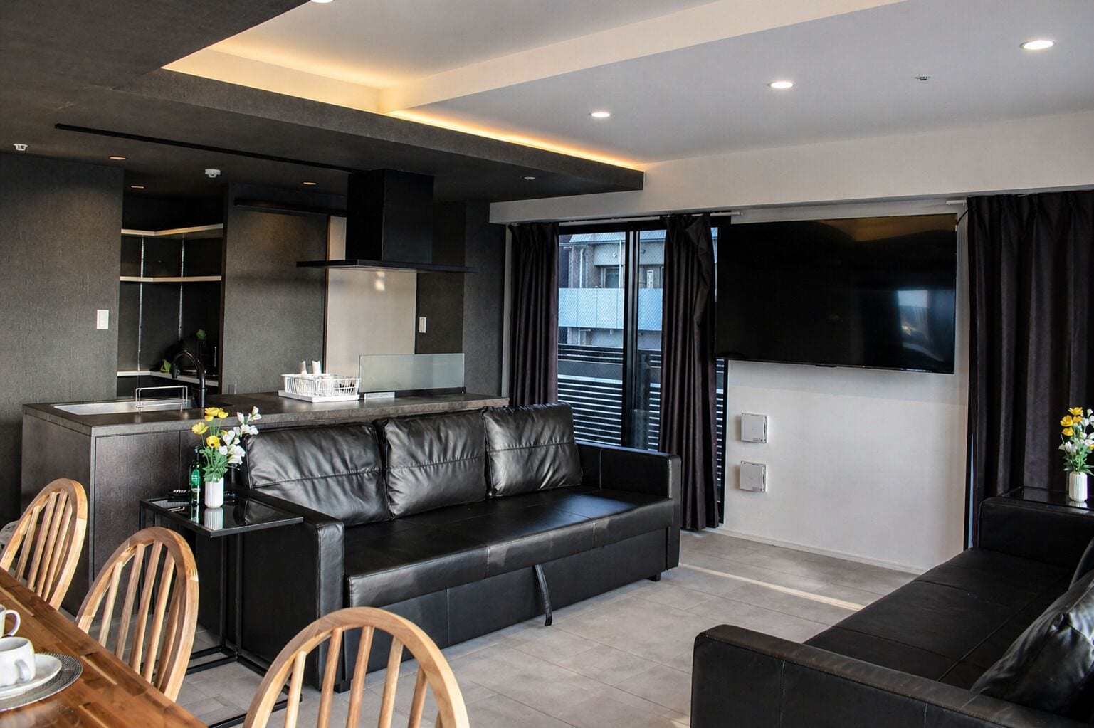 A modern living room with a sofa, dining table, and chairs, a kitchen area, and a large TV screen.