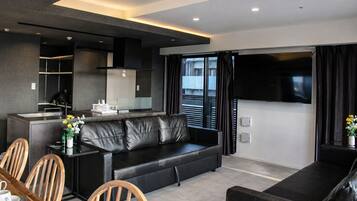 A modern living room with a sofa, dining table, and chairs, a kitchen area, and a large TV screen.