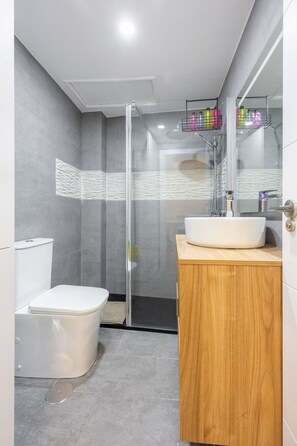 A modern bathroom with a glass shower enclosure, a white toilet, and a wooden vanity with a sink. - Stay in La Latina Stylish Apartment for Work and Study (Madrid)