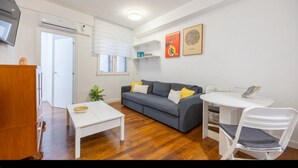 Un espacio habitable compacto con un sofá gris, una mesa de comedor blanca y una pequeña silla blanca. Hay un mueble de madera y un televisor montado en la pared.