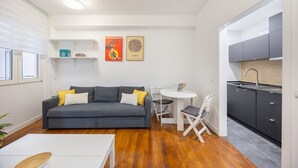 Un espacio habitable compacto con un sofá gris, una pequeña mesa de comedor y una cocina con armarios grises y encimera blanca.