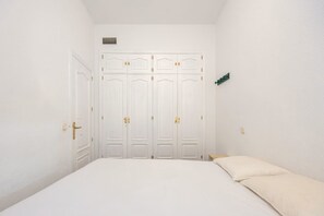 Quarto com cama, paredes brancas, guarda-roupa branco com puxadores dourados e uma pequena planta verde na parede.