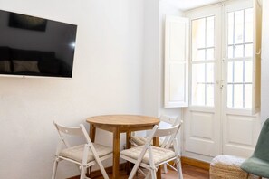 Quarto com uma mesa redonda de madeira, quatro cadeiras brancas, uma TV de tela plana fixada na parede e uma porta branca com painéis de vidro.