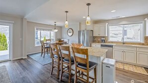 Une cuisine moderne avec un coin repas, comprenant un grand îlot central, des chaises en bois, des luminaires suspendus et une vue sur l’extérieur.