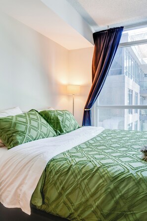 A bed with green bedding, a bedside lamp, and a window with curtains. - Downtown Toronto Studio Suite by the Water & FIFA (Toronto)