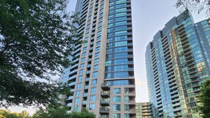 Un immeuble moderne de grande hauteur, doté de nombreux étages et de balcons, entouré d’arbres et bordé d’un trottoir dallé.