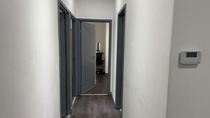 Un couloir étroit aux murs gris et au plancher en bois, qui mène à une pièce où se trouvent une chaise noire et un miroir.