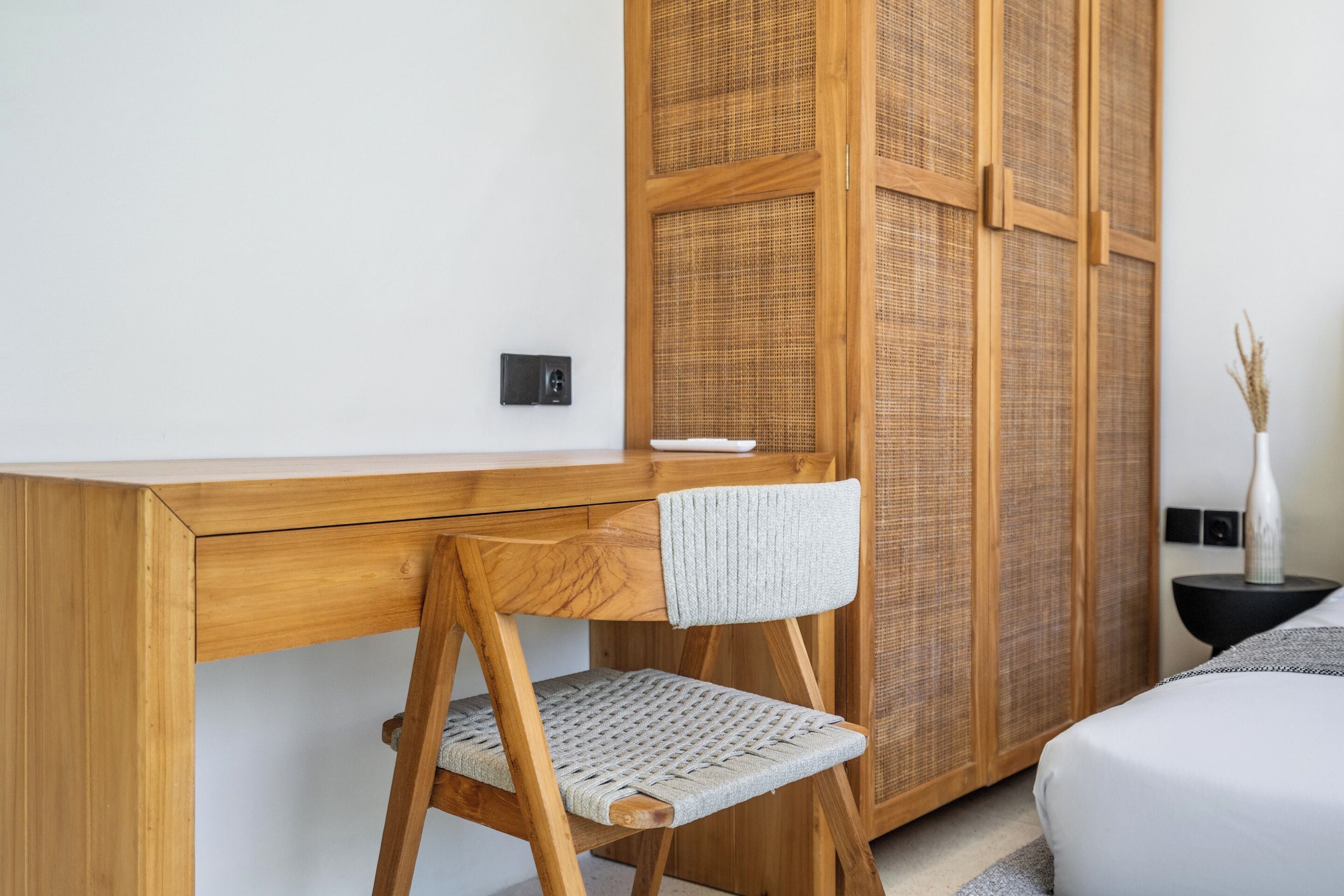 A wooden desk with a woven chair, a wardrobe with woven doors, and a bedside table with a vase.