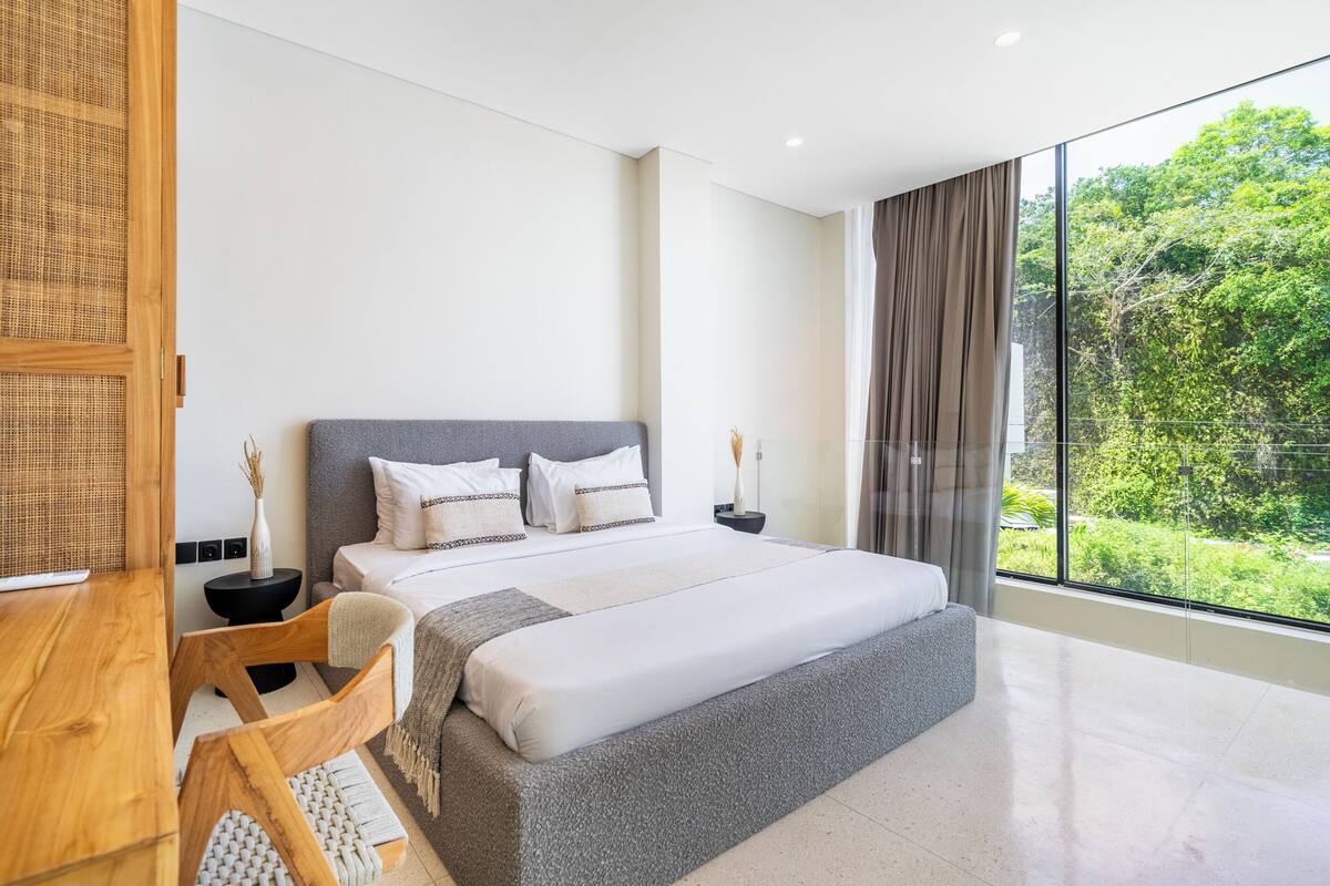 A modern bedroom with a large bed, a wooden desk, and a view of greenery outside.