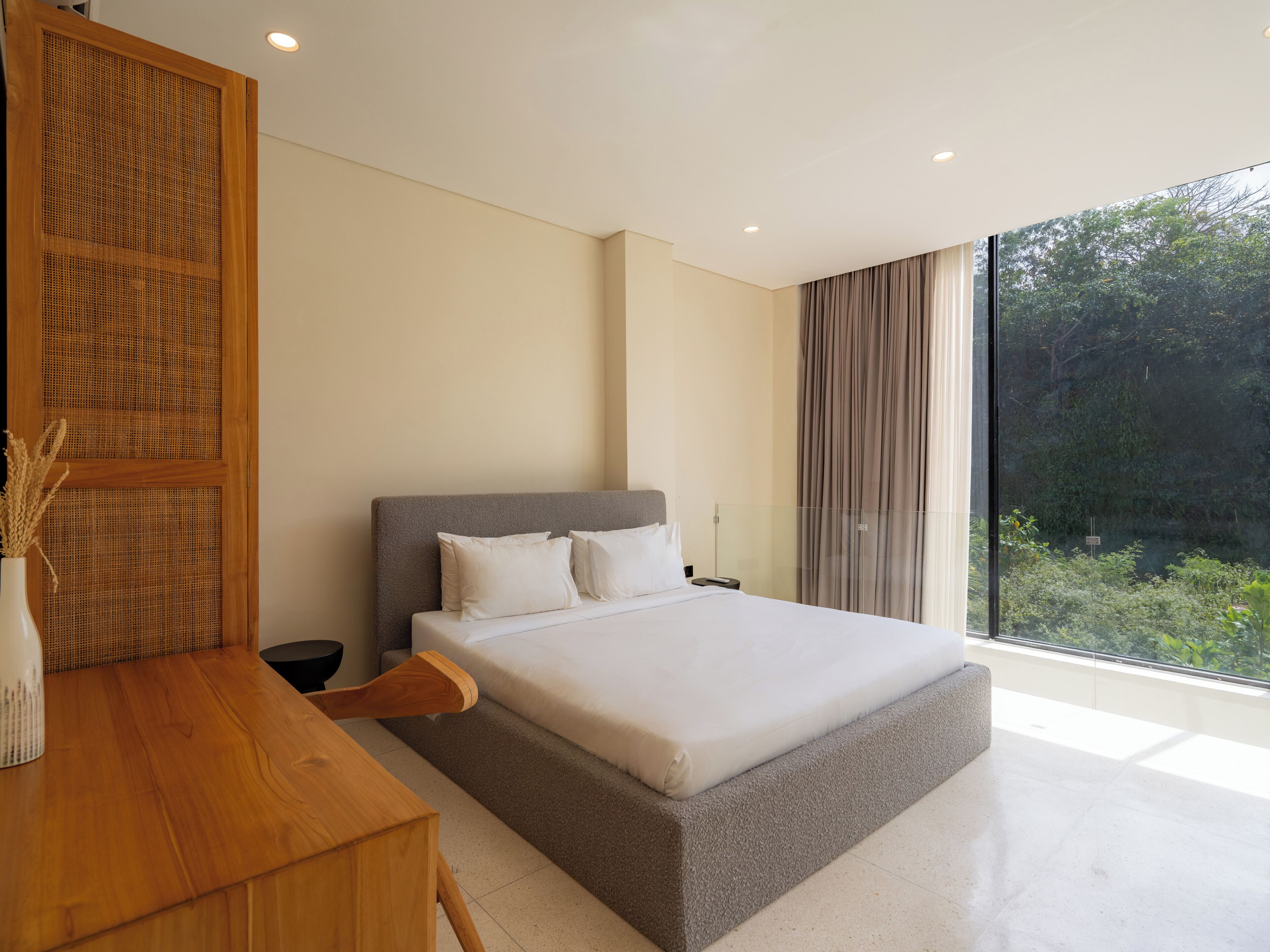 A modern bedroom with a large bed, a wooden desk, and a view of greenery through a large window.