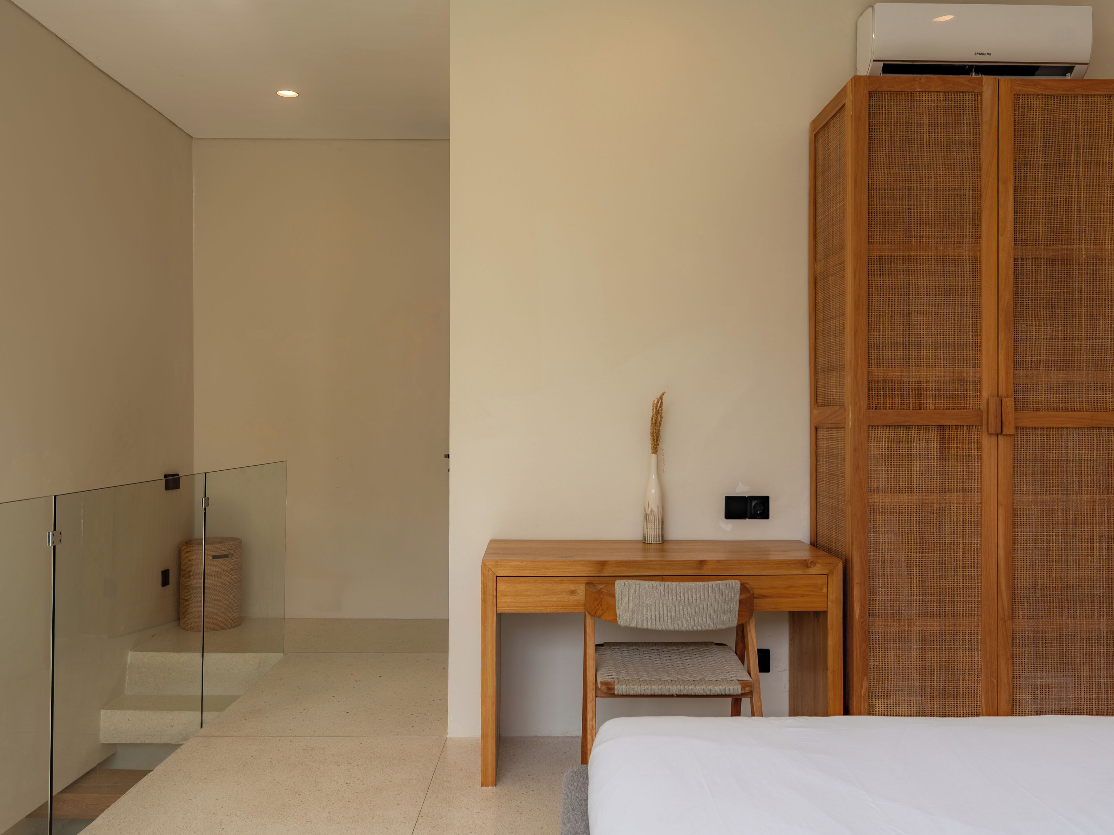 A bedroom with a bed, a wooden desk, a chair, a bedside table, a wall-mounted air conditioner, and a woven basket.