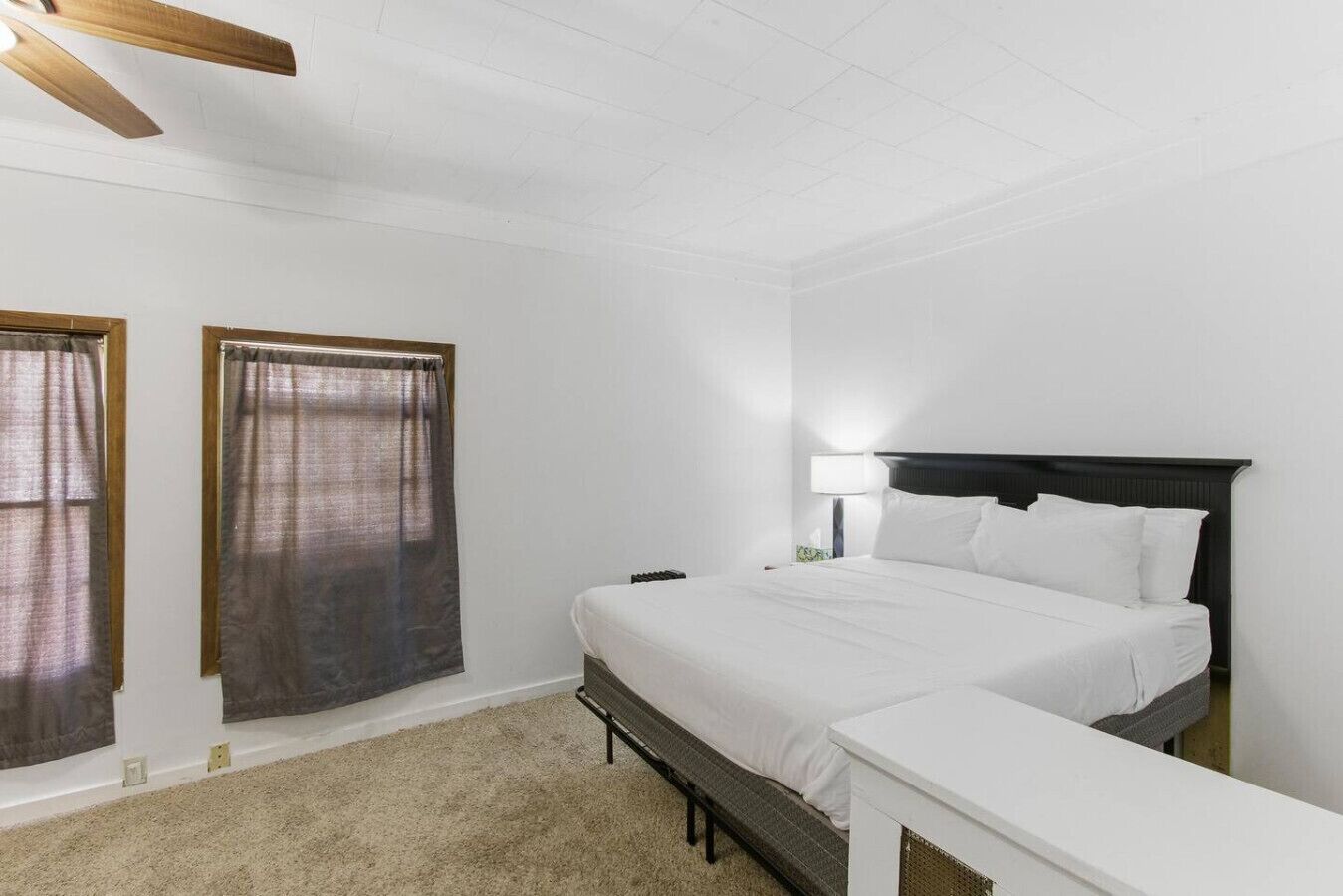 Un dormitorio con una cama, una mesita de noche, una lámpara y dos ventanas con cortinas.