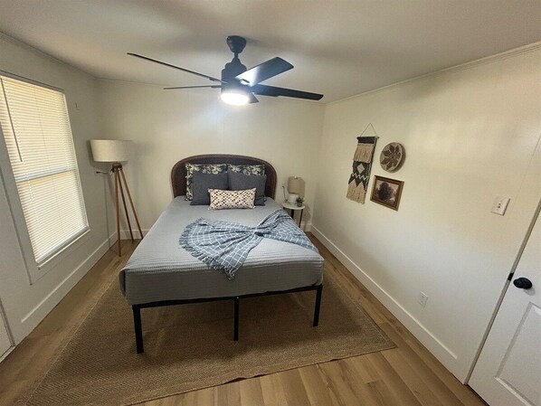 A bedroom with a bed, a ceiling fan, a window with blinds, a bedside table, and a wall clock.