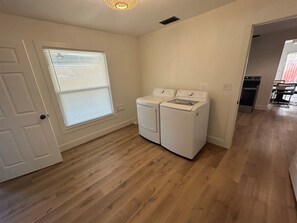 A laundry room with two white washing machines, wooden flooring, a door, and a window with blinds.