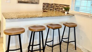 Una cocina con encimera de granito, cuatro taburetes de madera, una lámpara colgante, un cuadro en la pared y una ventana con persianas.