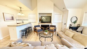 Una sala de estar moderna con un sofá, una mesa de centro y un televisor. Hay una zona de cocina con un taburete de bar y una chimenea.