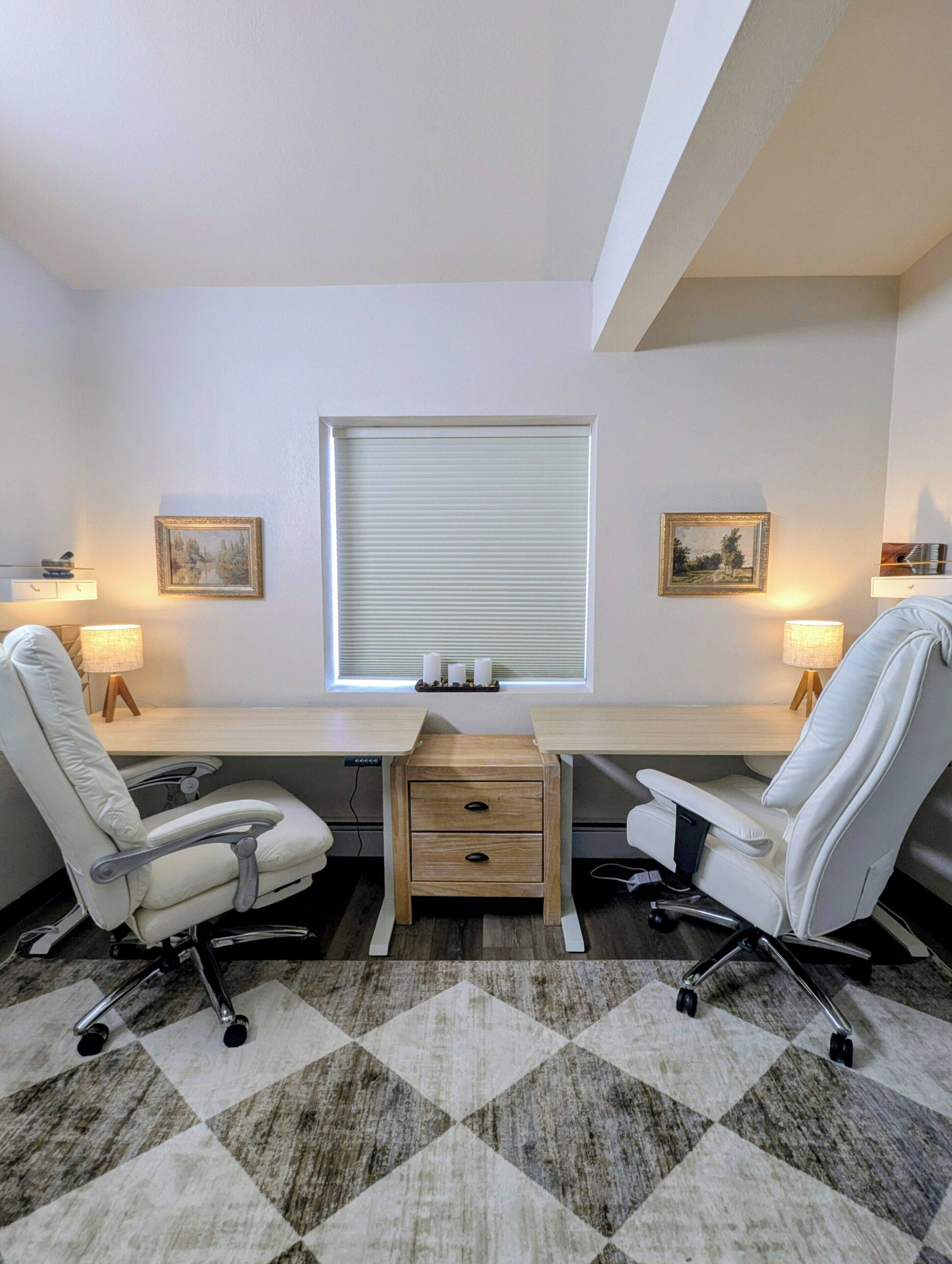 A modern office space with a desk, two office chairs, a small wooden drawer unit, and a window with blinds.