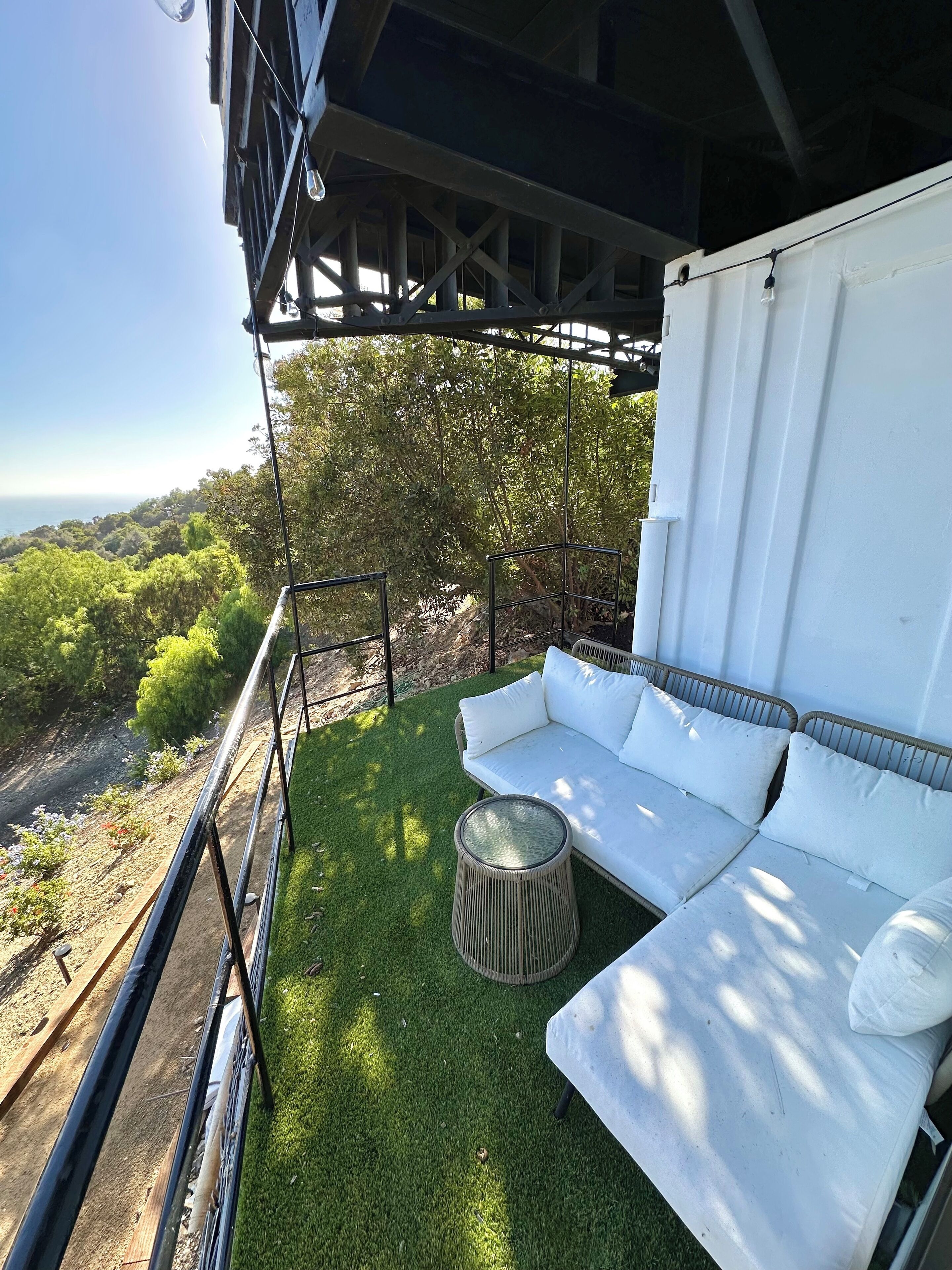 A balcony with a white sofa and a round wicker table, overlooking a lush green landscape and a body of water.