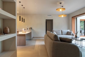 A modern interior with a sofa, pendant lights, a kitchen area, and a view through a large window.