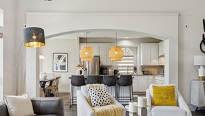 A modern living room with a sofa, armchair, and coffee table, leading to a kitchen with white cabinets and pendant lighting. - Spacious 6BR Villa for 14 · Home Theater, Game Loft & Gym · 10 Min to Universal (McKinney)