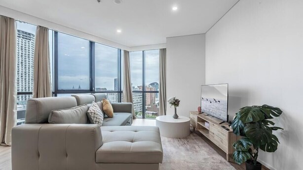 Uma sala de estar moderna com um sofá, uma televisão, uma mesa de centro redonda e uma janela grande com vista para a cidade.