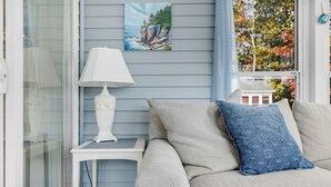 Une pièce avec un mur bleu clair, une lampe blanche posée sur une petite table, un canapé beige agrémenté d’un coussin bleu et un tableau représentant un paysage côtier.