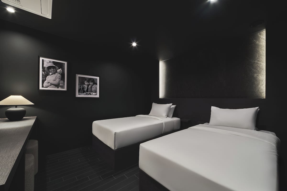 A modern hotel room with two single beds, a desk with a lamp, and a framed photograph on the wall.