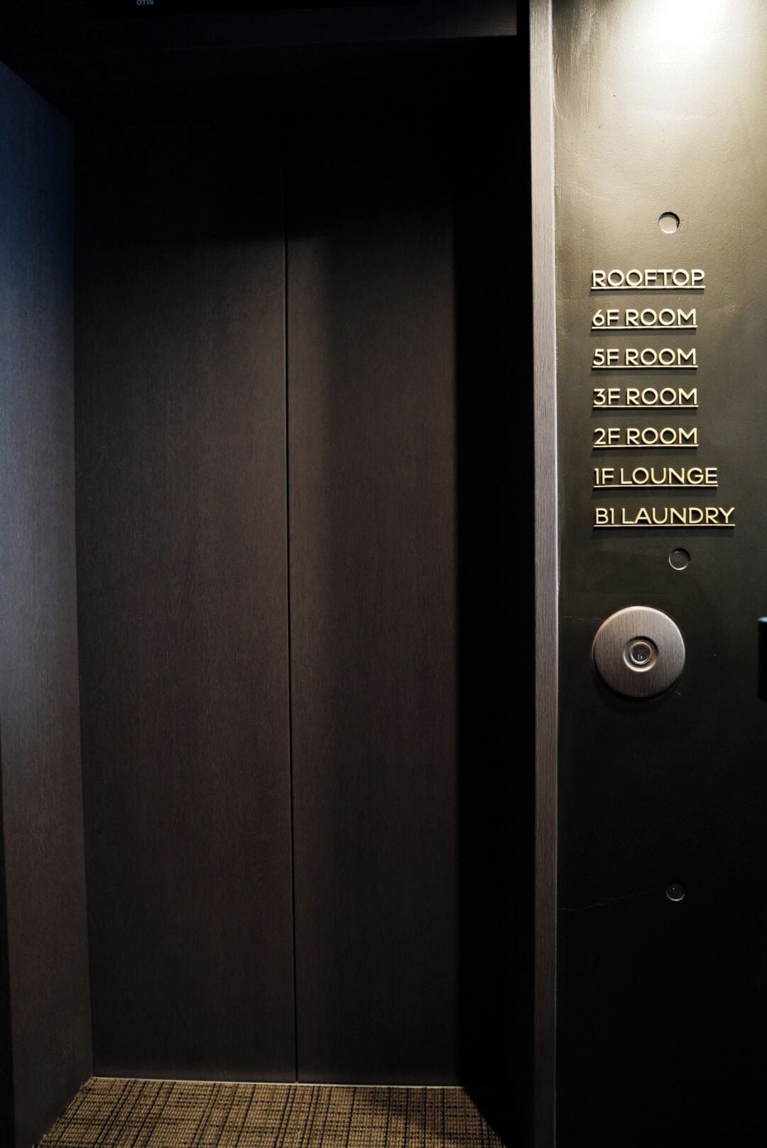 An elevator interior with a dark wooden door and a metallic wall with room labels including "6F ROOM," "5F ROOM," "3F ROOM," "2F ROOM," "1F LOUNGE," and "B1 LAUNDRY."