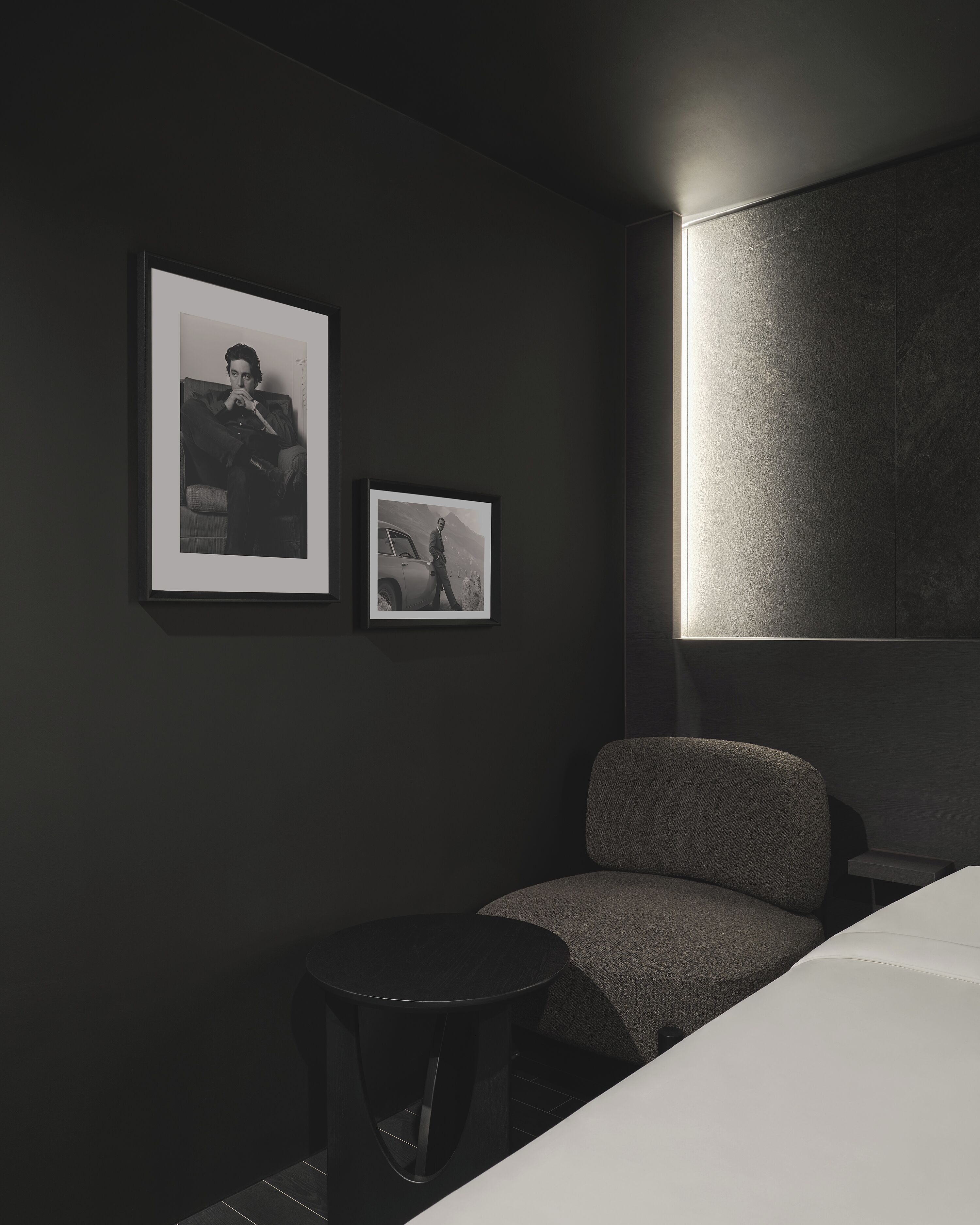 A dark-themed hotel room with a single grey armchair, a small round table, and two framed black and white photographs on the wall.