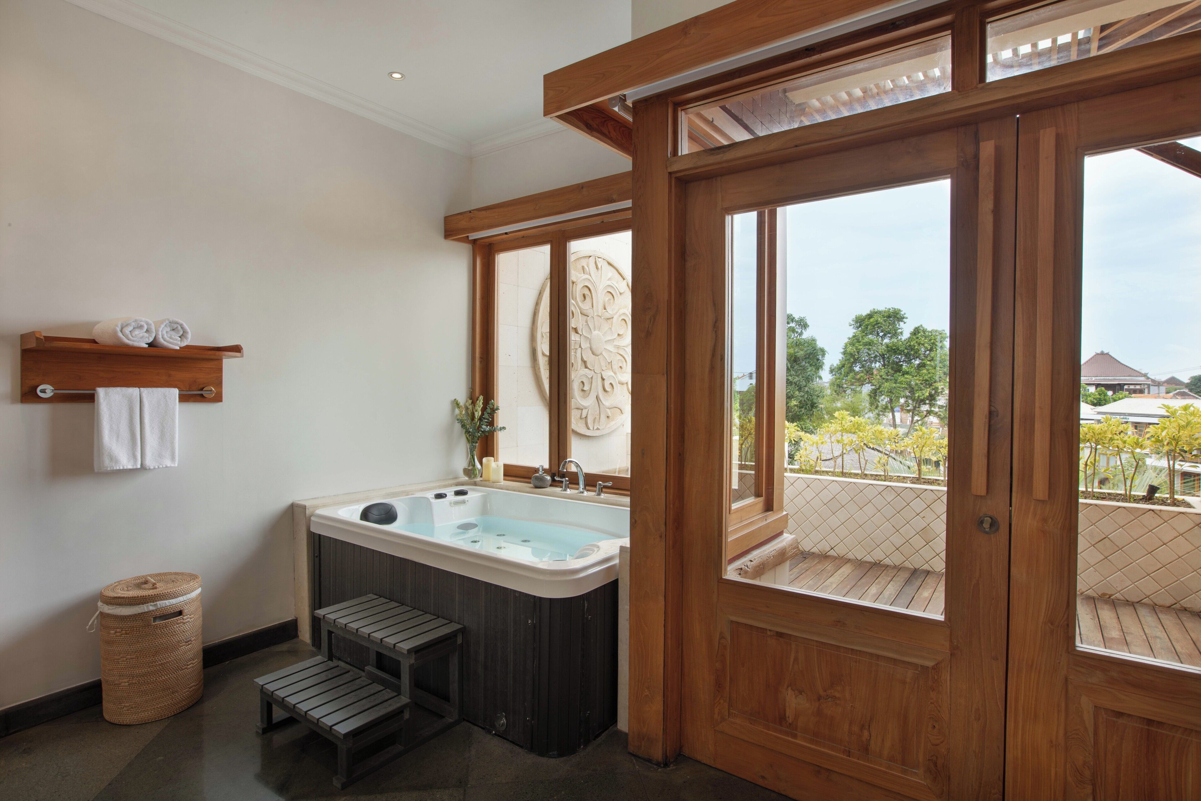 A bathroom with a wooden bathtub, a small stool, a towel rack, and a view through a wooden door.