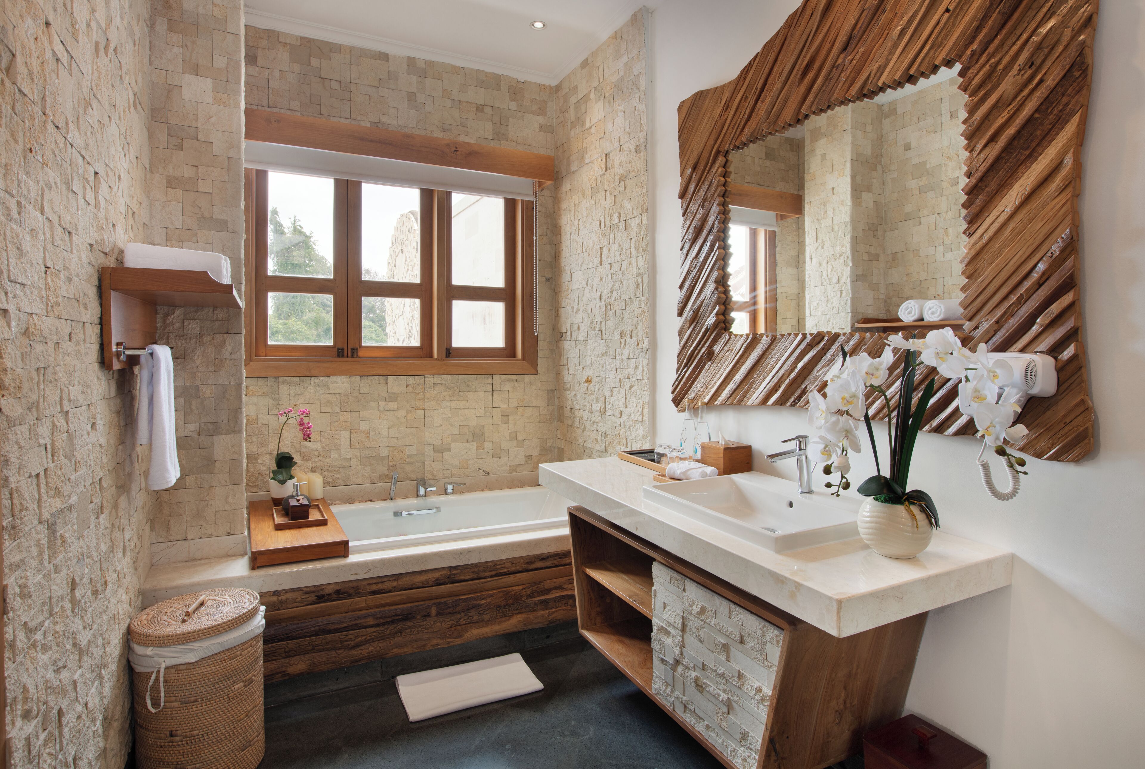 A modern bathroom with a stone wall, a large window, a wooden vanity, a bathtub, and a mirror with a wooden frame.