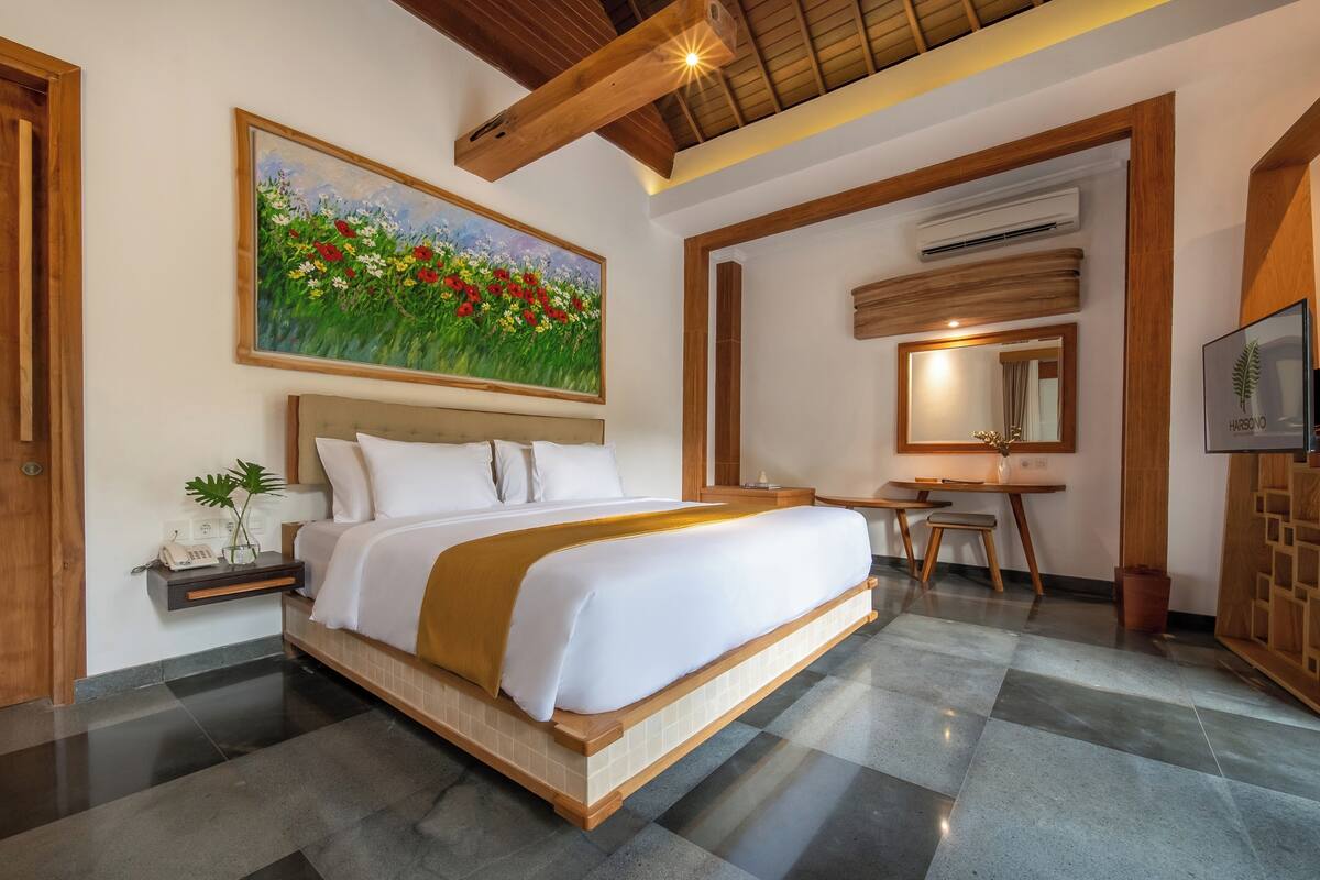 A hotel room with a large bed, a desk, a chair, a small table with a plant, and a painting of flowers on the wall.