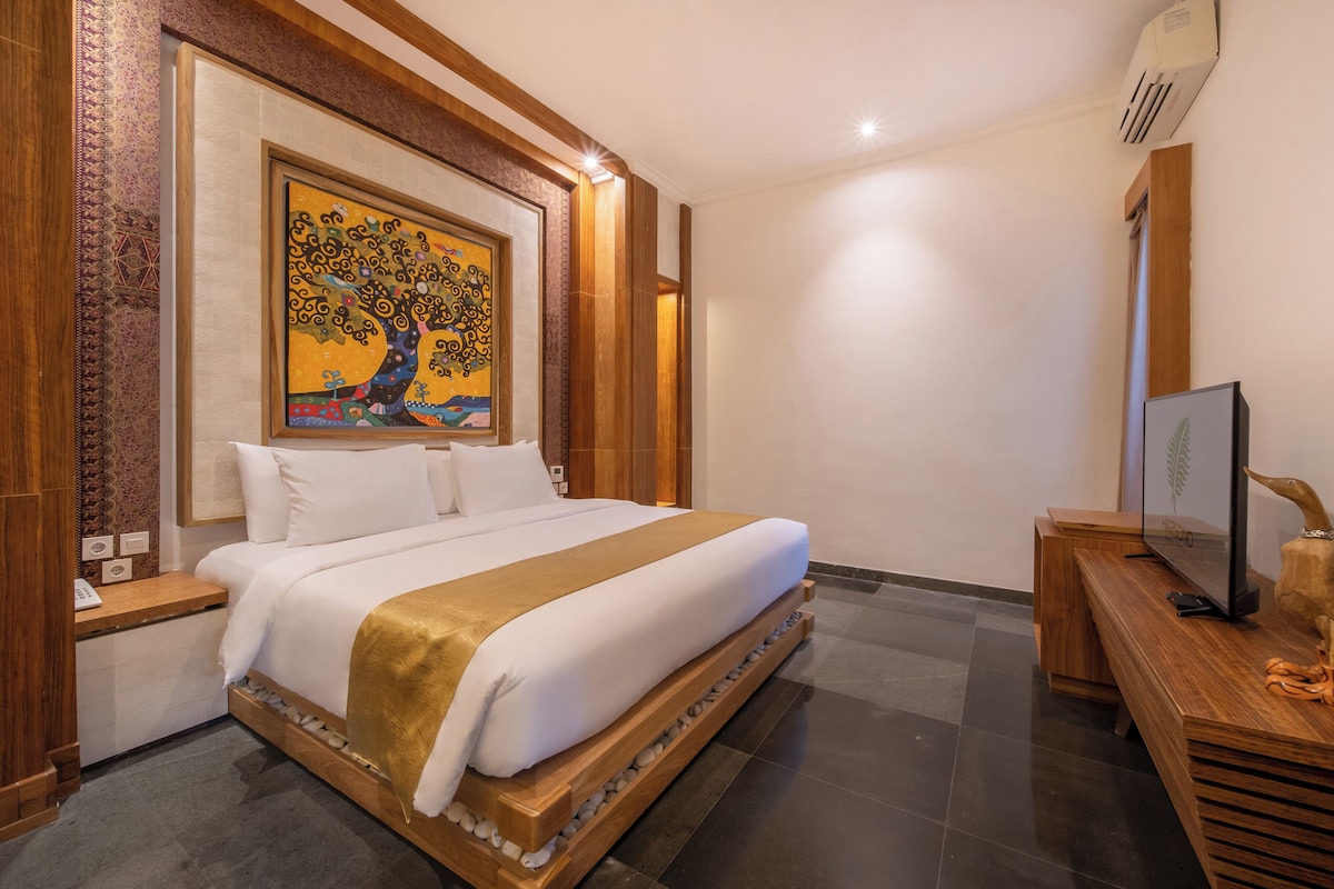 A bedroom with a large bed, a wooden headboard, a painting above the bed, a wooden nightstand, a television on a wooden stand, and a wall-mounted air conditioner.