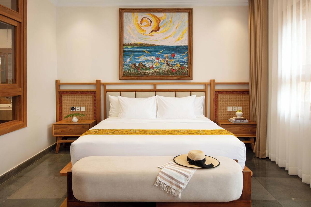 A hotel room with a large bed, wooden headboard, two bedside tables, a painting on the wall, and a window with curtains.