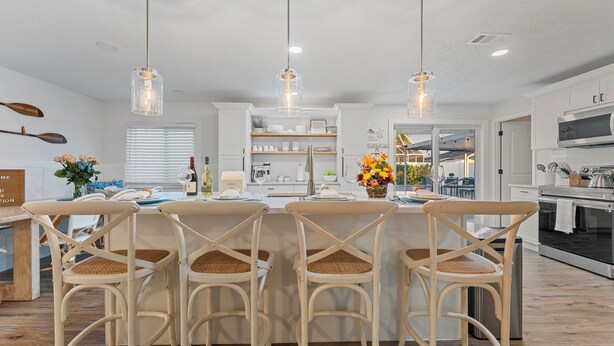 Una cocina moderna con armarios blancos, una isla central con taburetes altos, iluminación colgante y vistas al comedor.