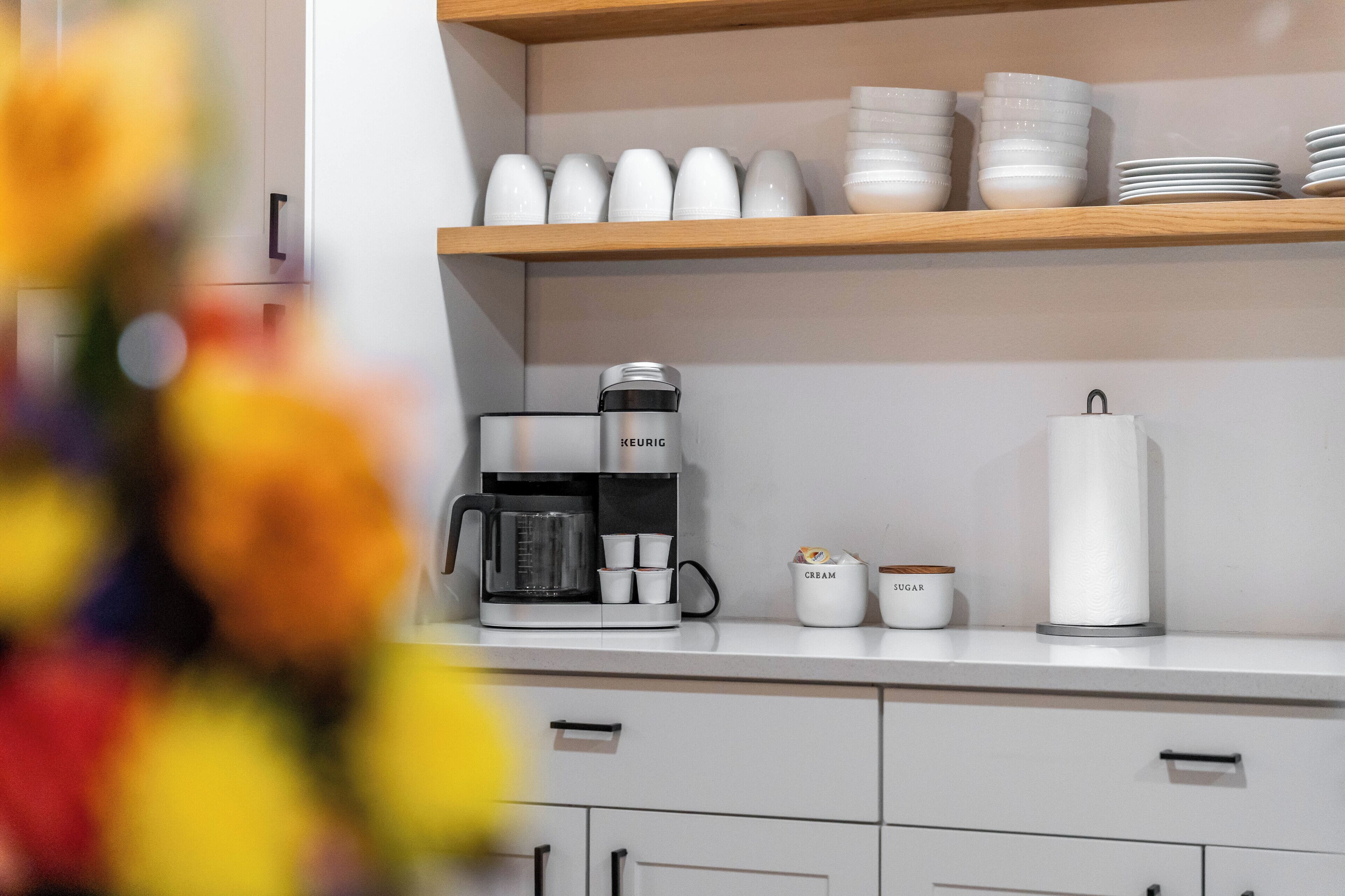 A kitchen with a coffee maker, a tea kettle, and storage jars labeled "CREAM" and "SUGAR" on a shelf.